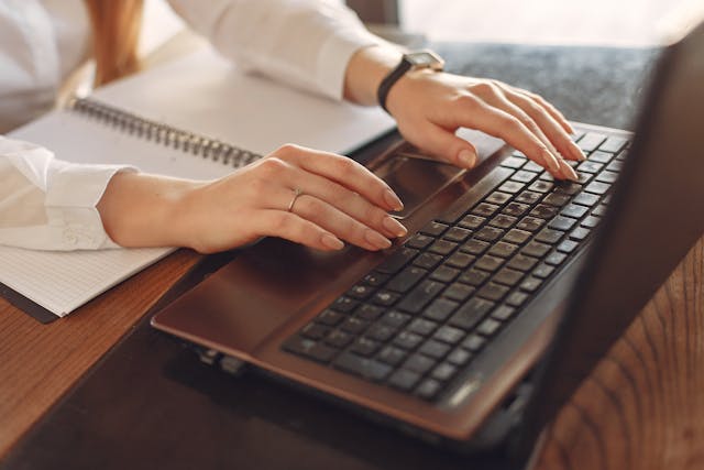 Eine Frau schreibt auf einem Laptop einen Blogbeitrag. Vor Ihr auf dem Tisch liegt ein aufgeschlagenes Notizbuch.